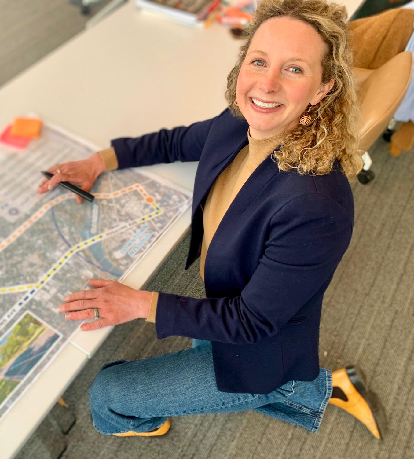 A white woman with curly hair wearing a blue blazer looking up from a table with a map on it.