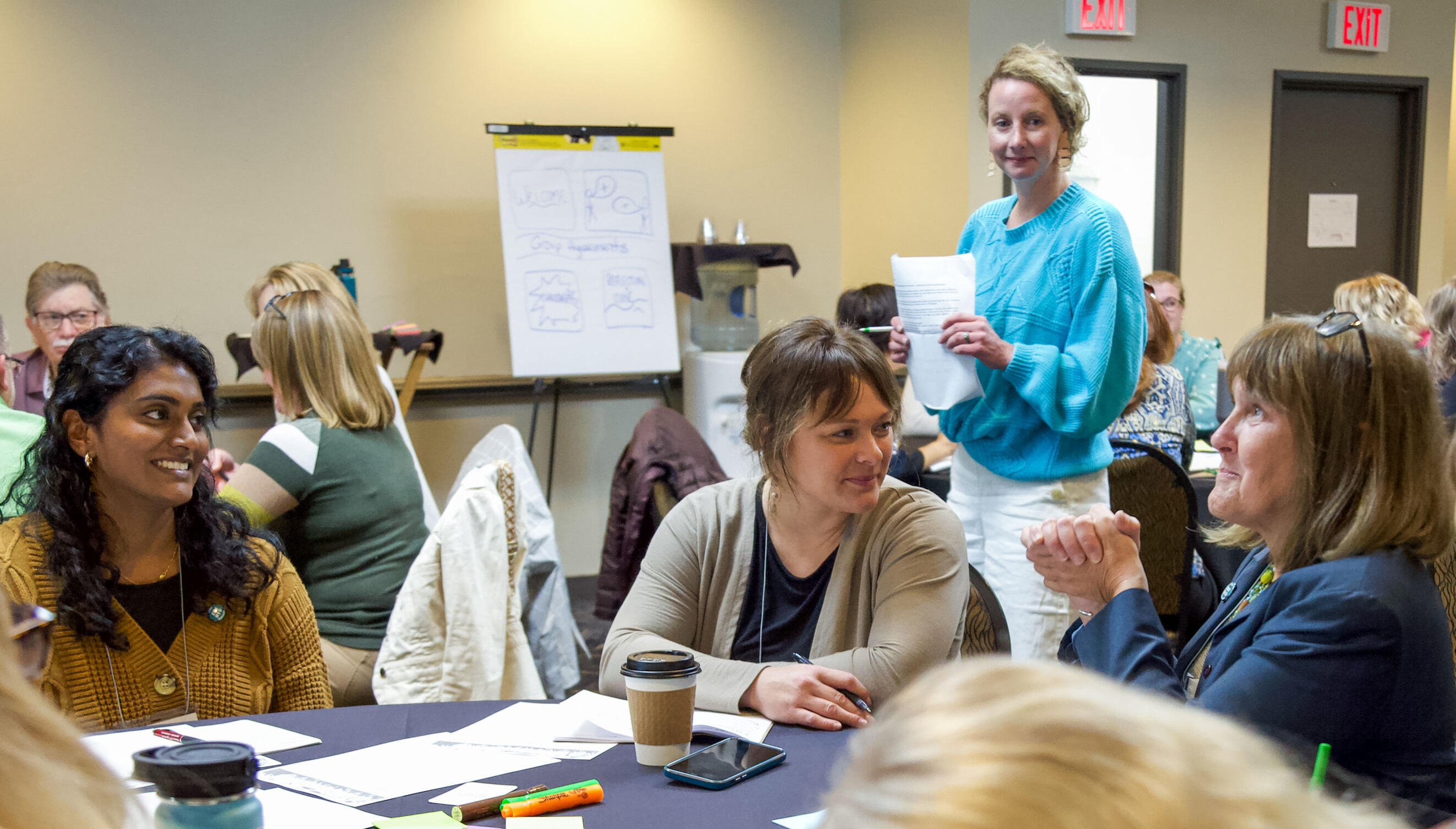 Three women sitting at a table in a conference room setting. One woman in a turquoise sweater is standing behind them, holding a paper and looking on with a smile.